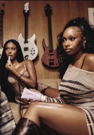Two African-American women indoors in chic dresses and boots, one applying moisturizer, with guitars in the background, warm lighting.
