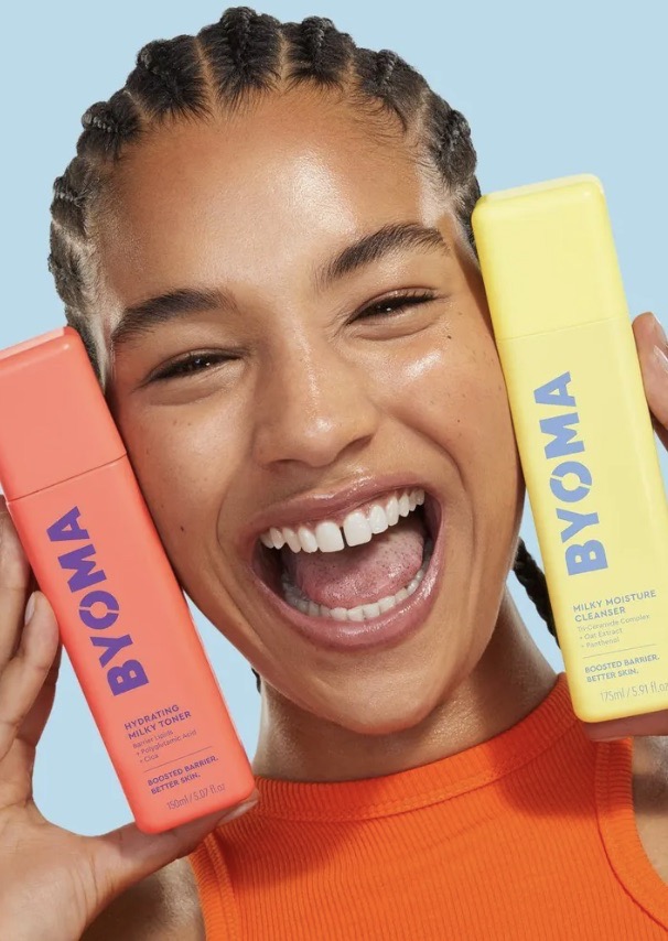 Close-up shot of an adult female holding BYOMA hydrating milky toner and cleanser, smiling brightly with a blue background.