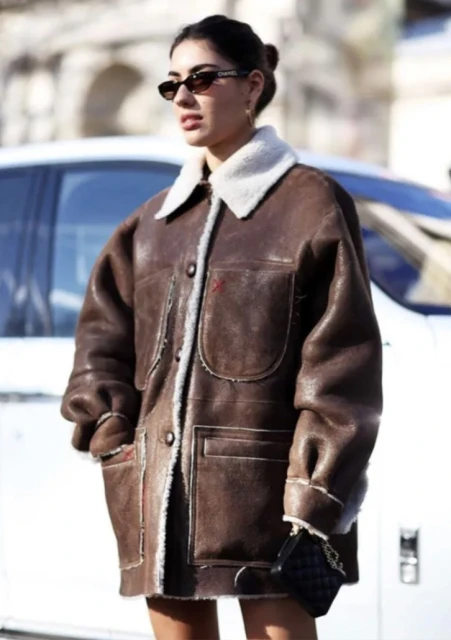 Fashionable adult woman with sunglasses and oversized brown sheepskin coat poses outdoors, holding a black quilted purse.