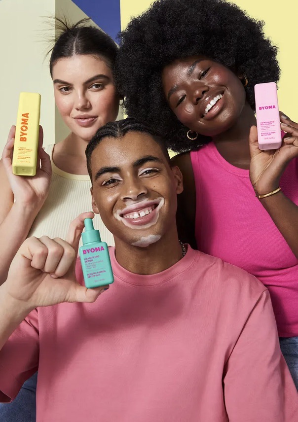 Three diverse models showcase BYOMA skincare products in a bright studio setting. The shot highlights their radiant skin and product packaging.