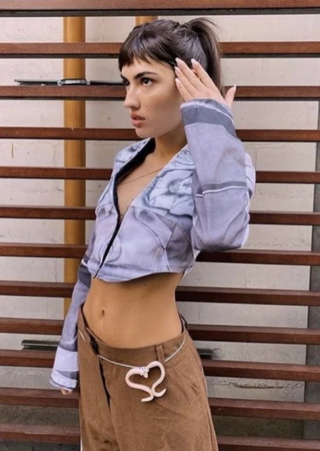 A woman with dark hair, wearing a cropped blouse and brown pants with a heart belt, poses indoors against a background of brown-painted steps.