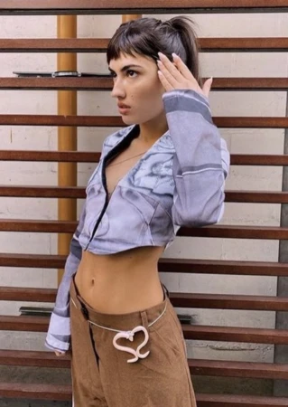 A woman with dark hair, wearing a cropped blouse and brown pants with a heart belt, poses indoors against a background of brown-painted steps.