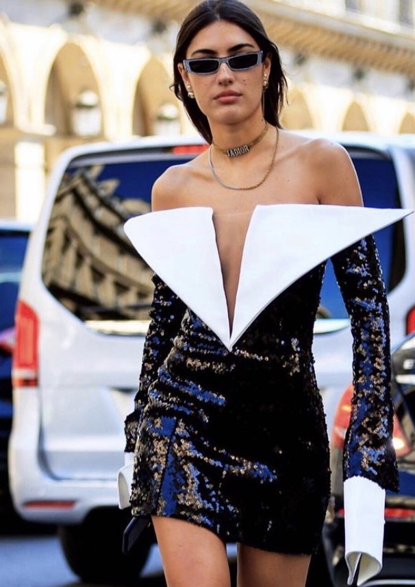 Brunette woman in sequined mini dress with white collar and cuffs stands on a city street with white van in the background.