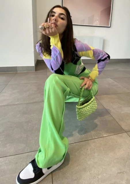 Brunette woman posing in vibrant lime pants, argyle sweater, and sneakers. Indoor shot in front of neutral wall and radiator.