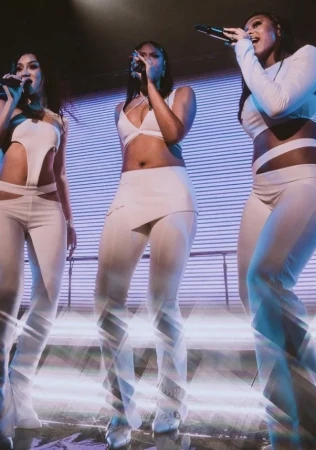 Three women sing on stage in matching outfits with microphones, illuminated by blue-toned stage lighting and a digital backdrop screen.