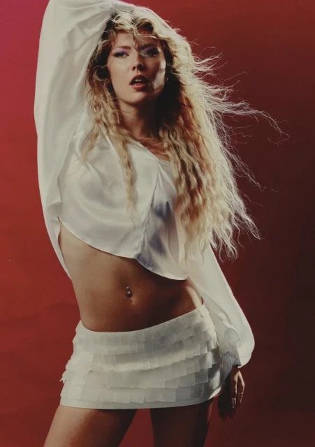 Blonde woman in white dress poses against red background. Close-up portrait with wind-blown hair and fashion style, indoor studio shot.