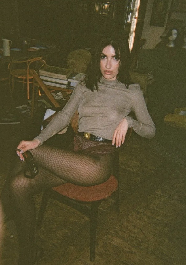 Brunette woman in long-sleeved top and patterned shorts sits on a red chair in an indoor setting, holding sunglasses.