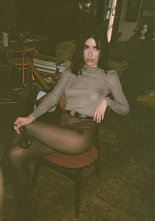 Brunette woman in long-sleeved top and patterned shorts sits on a red chair in an indoor setting, holding sunglasses.