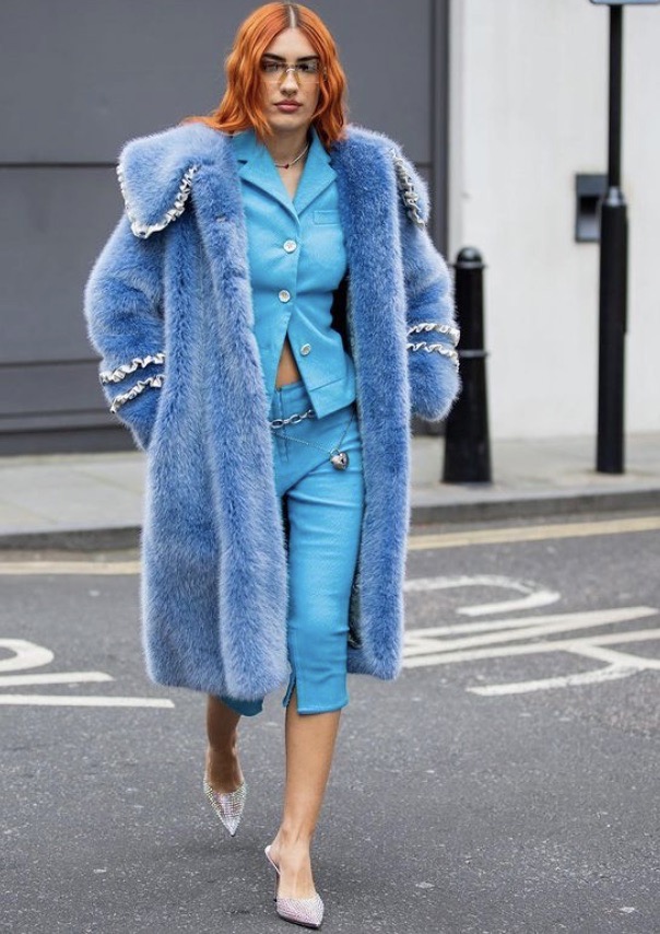 A woman in a blue faux fur coat and matching suit walks on a city street, showcasing a bold fashion statement with designer heels.