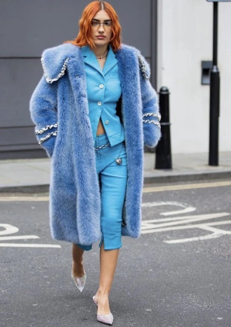 A woman in a blue faux fur coat and matching suit walks on a city street, showcasing a bold fashion statement with designer heels.