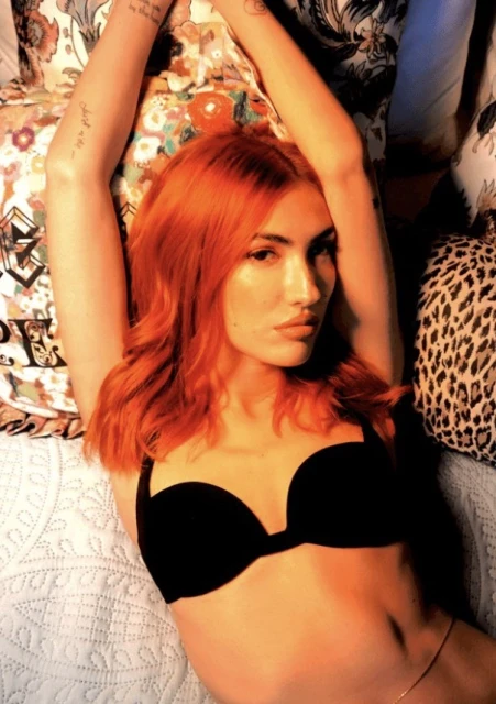 Attractive woman with red hair lies on a bed with black bra, in a studio setting. Floral and animal print pillows surround her.