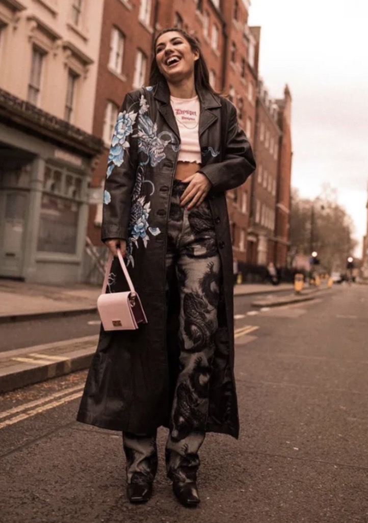 Smiling woman wearing embroidered black leather coat and patterned jeans holding a pink handbag on a city street.