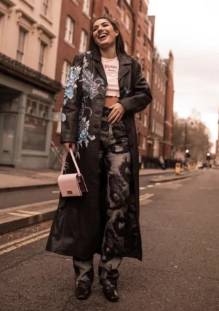Smiling woman wearing embroidered black leather coat and patterned jeans holding a pink handbag on a city street.