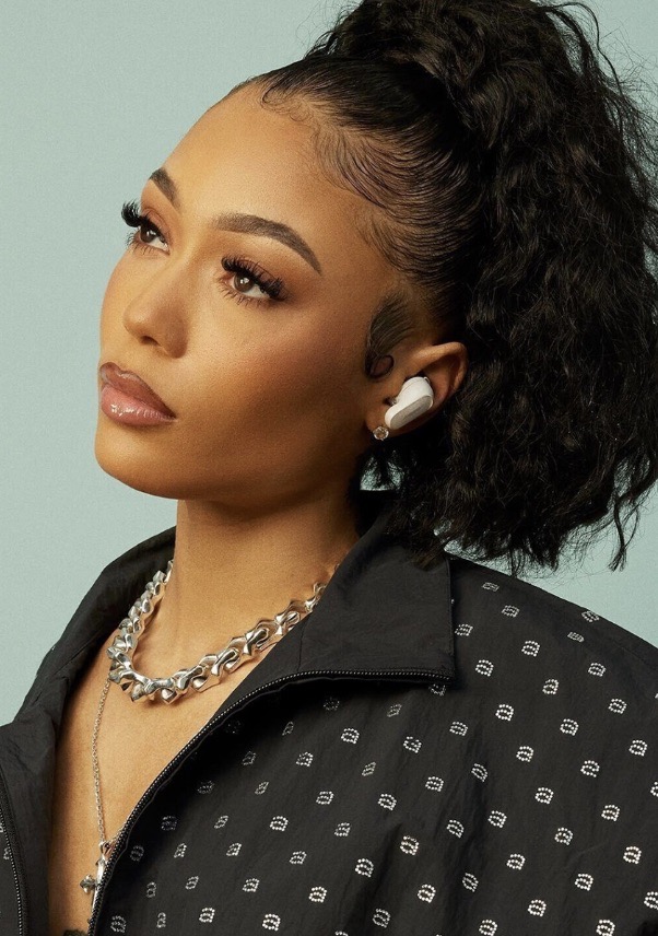 Close-up shot of a woman with dark curly hair in a high ponytail wearing a jacket and jewelry looking up and to the left.