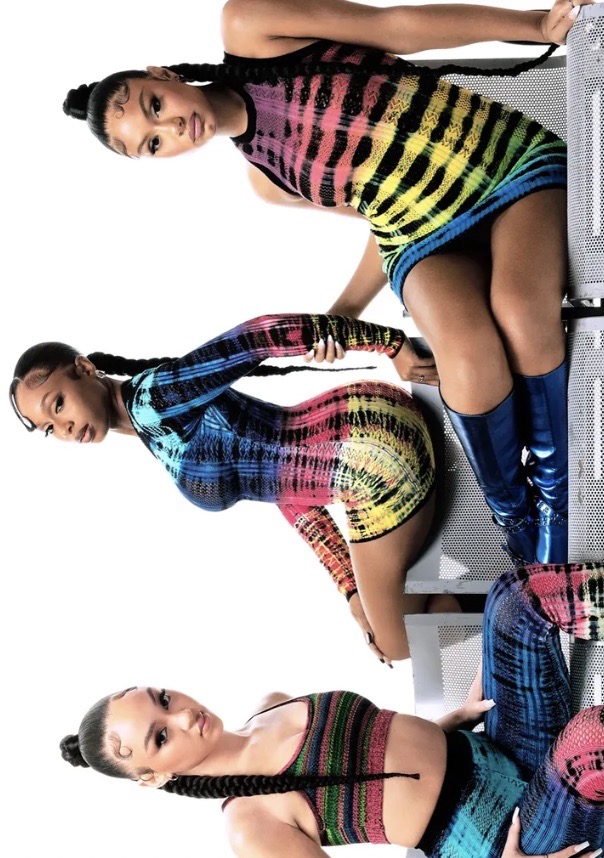 Three women pose in a studio wearing colorful tie-dye mini-dresses. Each has long braids and stands near metallic grating.
