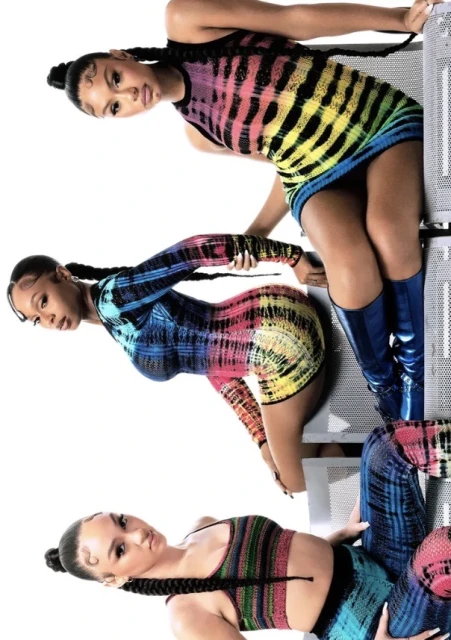Three women pose in a studio wearing colorful tie-dye mini-dresses. Each has long braids and stands near metallic grating.