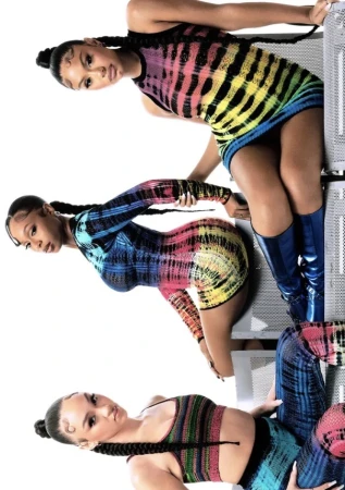 Three women pose in a studio wearing colorful tie-dye mini-dresses. Each has long braids and stands near metallic grating.