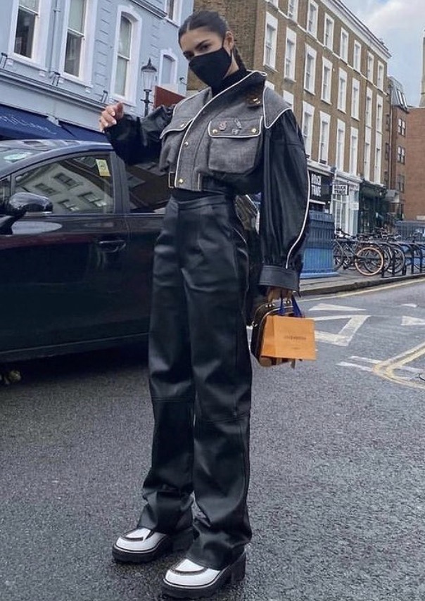 A woman in a fashionable grey jacket, black leather pants, and platform shoes walks on a city street carrying a Louis Vuitton bag.