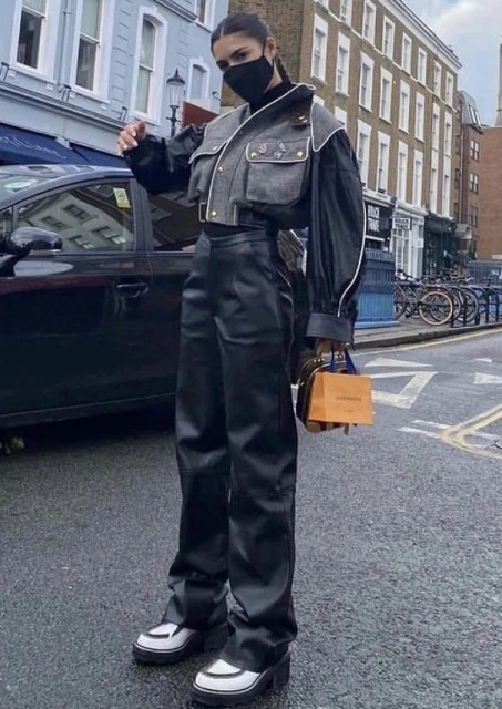 A woman in a fashionable grey jacket, black leather pants, and platform shoes walks on a city street carrying a Louis Vuitton bag.