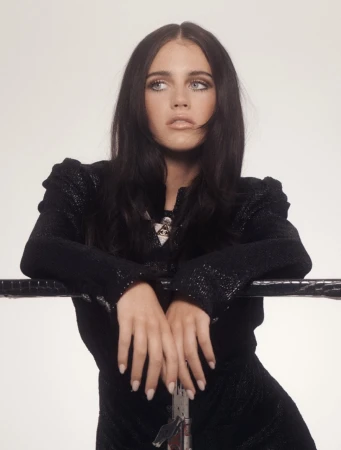 A dark-haired woman leaning against a black railing wearing a black jacket against a plain background