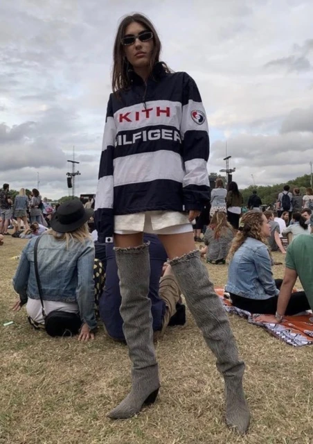 A full-shot of a woman in a striped jacket and thigh-high boots at an outdoor event, with a large crowd in the background.