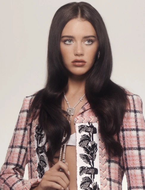 Portrait of a woman with long brunette hair wearing a pink plaid jacket, necklaces, and earrings in a studio setting.