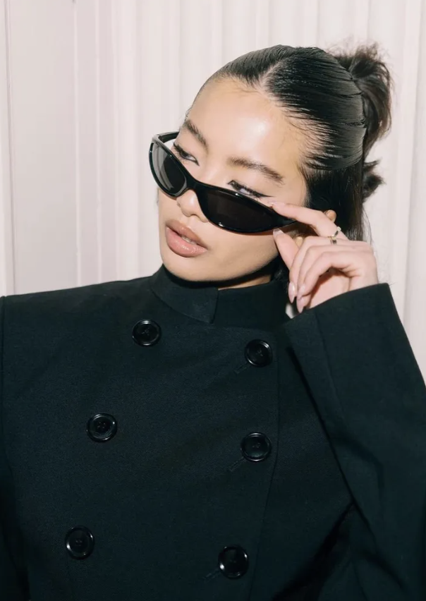 Asian adult female models sunglasses, wearing a black jacket, in a studio with a neutral-toned backdrop, beauty shot, eye-level.