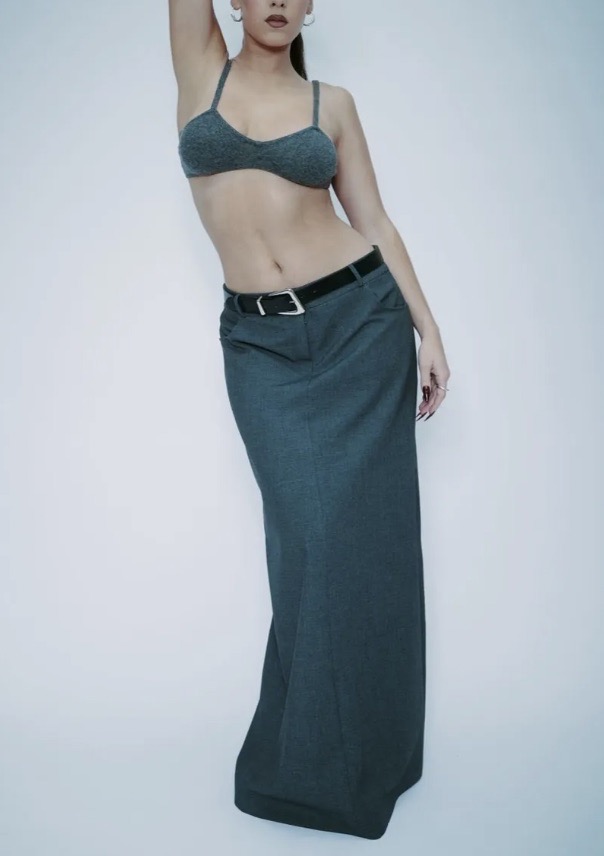Woman poses in a knit bra top and long skirt with a belt against a white background in a fashion forward editorial style. 
