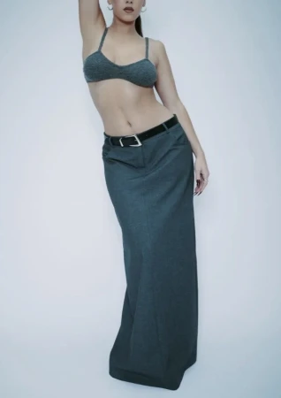 Woman poses in a knit bra top and long skirt with a belt against a white background in a fashion forward editorial style. 