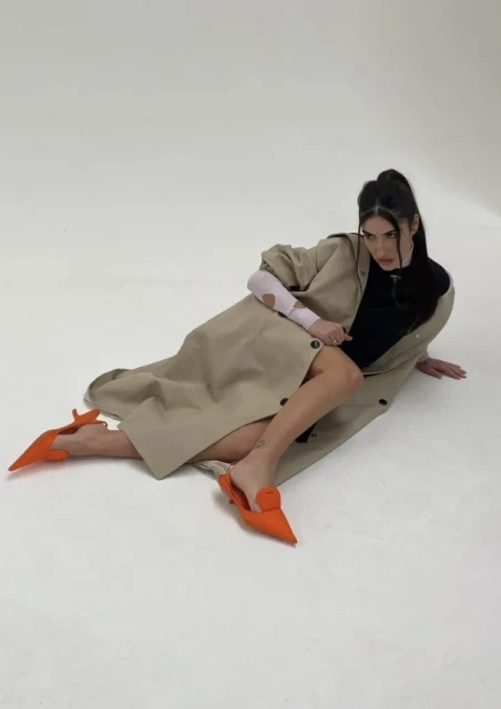 A woman lies on a white surface in orange shoes and a tan coat, gazing directly at the camera in a fashion pose, studio lighting.