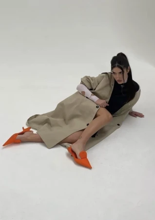 A woman lies on a white surface in orange shoes and a tan coat, gazing directly at the camera in a fashion pose, studio lighting.