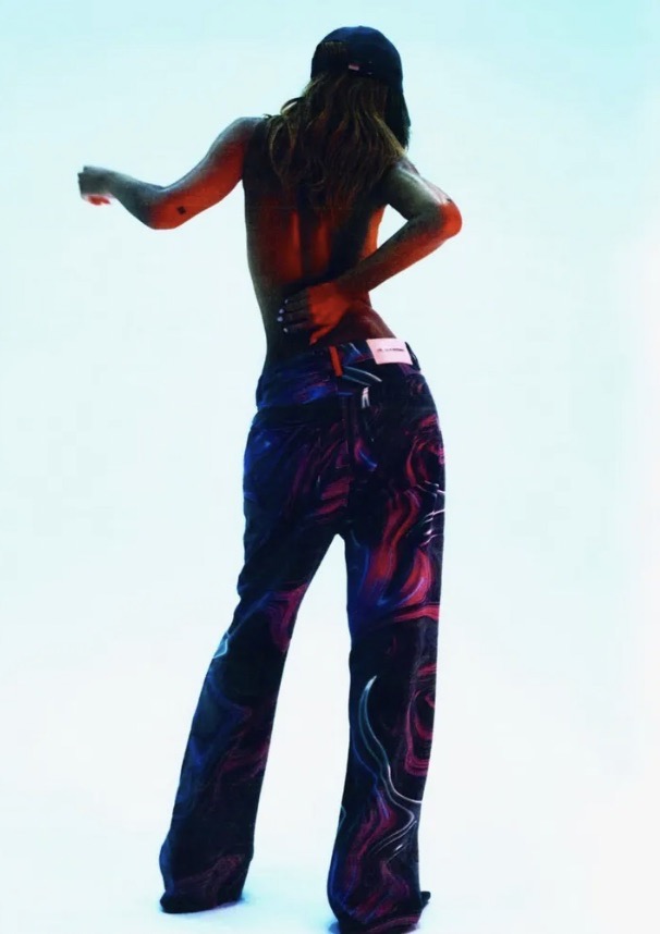 Back view of a slender person with long brown hair wearing a black baseball cap and printed jeans against a light blue background.