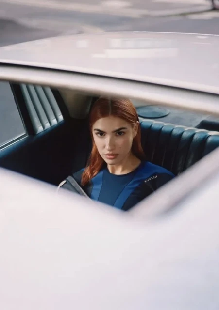 A woman with auburn hair in the back seat of a white car, wearing a blue athletic top and looking at the camera with a serious expression.