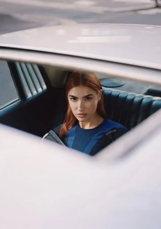A woman with auburn hair in the back seat of a white car, wearing a blue athletic top and looking at the camera with a serious expression.