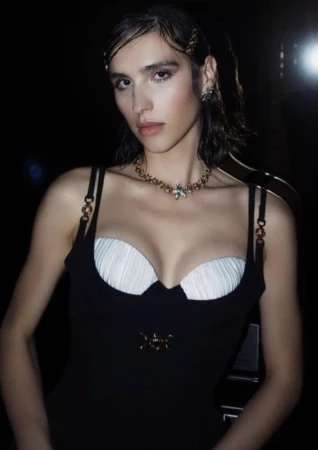 A woman in a black dress with white bustier styling wears a gold statement necklace and hair clips in a dimly lit setting.