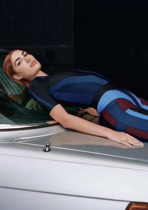 Adult woman lying on the hood of a silver car in a colorful bodysuit, gazing into the camera with a neutral facial expression.