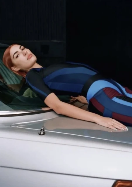 Adult woman lying on the hood of a silver car in a colorful bodysuit, gazing into the camera with a neutral facial expression.