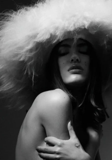 Monochrome portrait of a woman with closed eyes and a large, fluffy hat, her arms crossed and a tattoo visible on her hand.