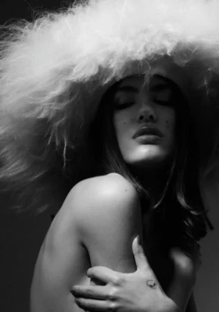 Monochrome portrait of a woman with closed eyes and a large, fluffy hat, her arms crossed and a tattoo visible on her hand.