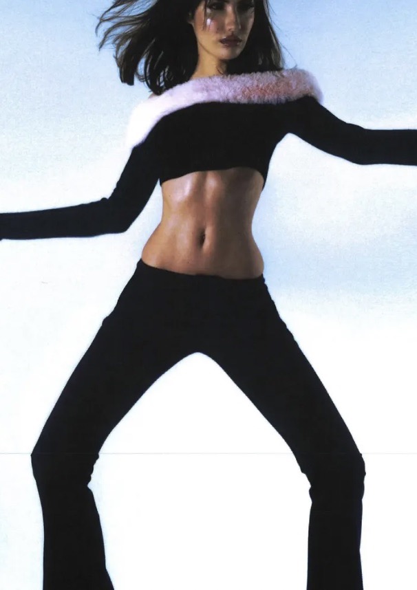 Fashion model in a black two-piece outfit with fur trim striking a wide-legged pose against a bright white background.