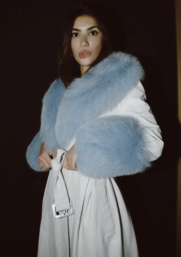 Woman in a tan trench coat with baby blue fur trim posing in a dimly lit indoor environment. Fur trim is around the neck and sleeves.
