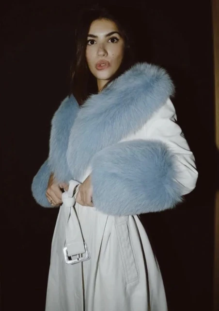 Woman in a tan trench coat with baby blue fur trim posing in a dimly lit indoor environment. Fur trim is around the neck and sleeves.