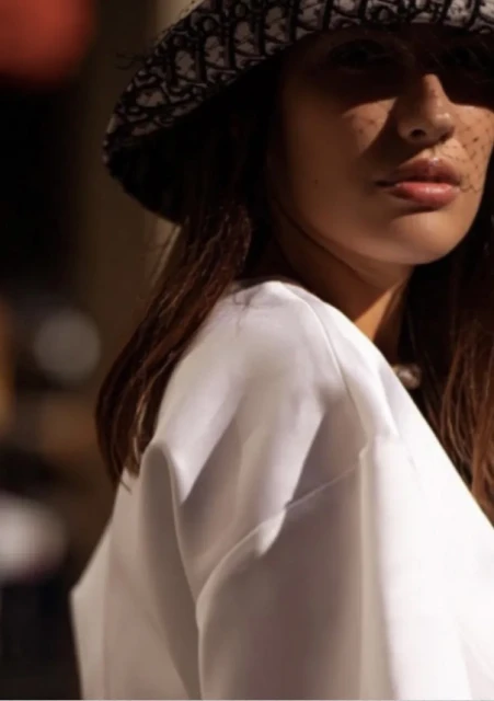 Close-up of a fashionable adult woman with freckles wearing a patterned bucket hat and white top, enjoying soft, directional sunlight.