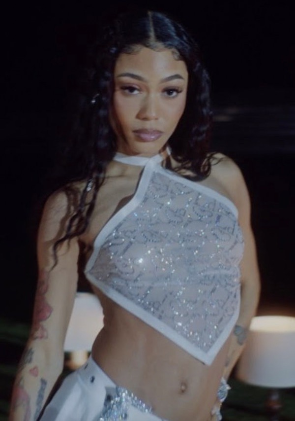Close-up of a young adult woman with long, dark hair, wearing a white halter top with silver embellishments, in a dimly lit environment.