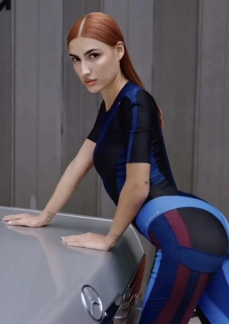 Attractive woman with auburn hair leans on the hood of a silver car wearing a navy and blue catsuit in a studio setting.