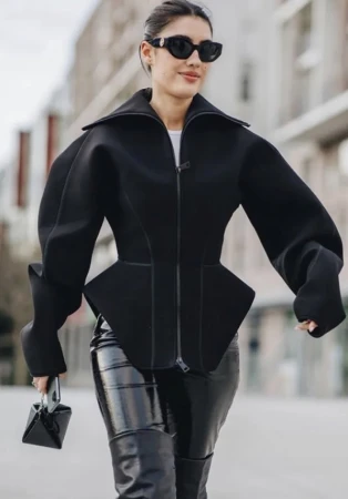 Model wearing black sunglasses, structured zip jacket, and shiny trousers walking outdoors.