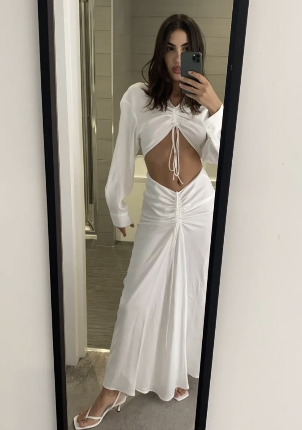 Woman in mirror selfie wearing a long-sleeved white dress with cutouts, accessorized with heeled sandals in an indoor setting.