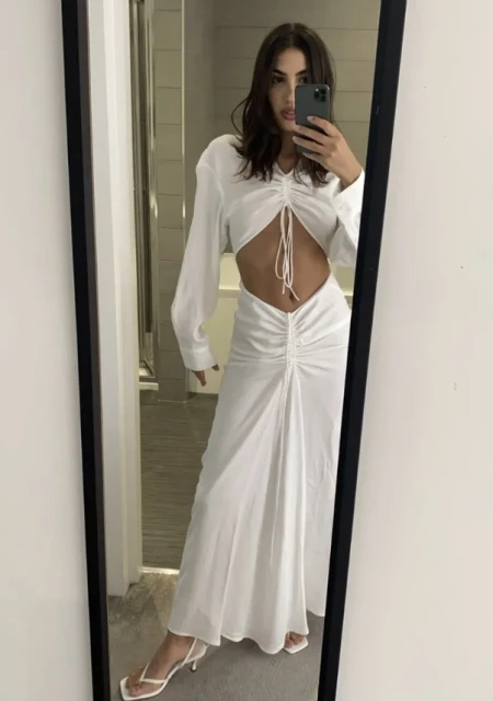 Woman in mirror selfie wearing a long-sleeved white dress with cutouts, accessorized with heeled sandals in an indoor setting.