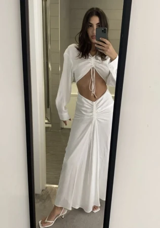 Woman in mirror selfie wearing a long-sleeved white dress with cutouts, accessorized with heeled sandals in an indoor setting.
