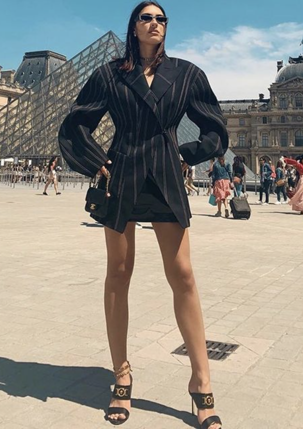 Woman in a blazer dress stands in front of the Louvre Pyramid in Paris, wearing sunglasses and heels with a small black purse.